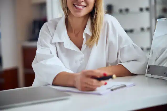 A woman in a white blouse is sitting at a desk, smiling. She holds a pen in her right hand, which rests on a clipboard with a piece of paper. A closed laptop sits on the desk beside her. The background is a blurry office space.