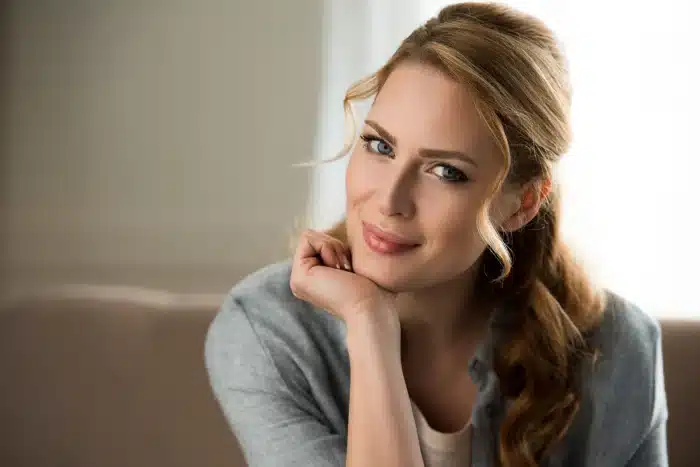 A woman with light brown hair, styled in loose waves, smiles softly while resting her chin on her hand. She is wearing a light blue buttoned shirt and is seated indoors in front of a blurred background. The lighting is soft and natural.
