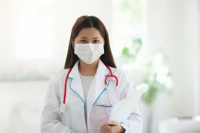 A female doctor with long brown hair stands indoors, wearing a white coat and a face mask. She holds a folder and has a red stethoscope around her neck. The background is bright and blurred, suggesting a clinical setting.