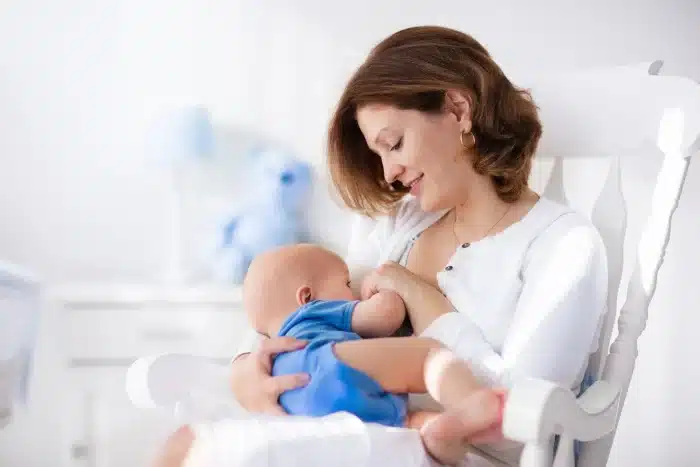 A woman with short, brown hair is sitting on a white rocking chair, breastfeeding a baby dressed in a blue onesie. The background features a white dresser and a plush teddy bear, creating a calm, nurturing environment.