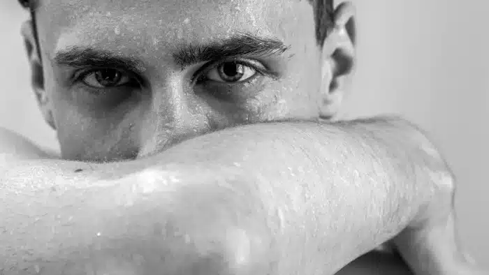 Black and white close-up of a person's face. They have intense eyes and are looking directly at the camera. Their arm is raised, covering the lower part of their face, and their skin is glistening with moisture.