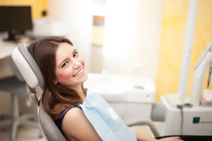 A young woman with brown hair is sitting in a dental chair, smiling at the camera. She is wearing a blue dental bib. The background shows dental equipment and a sink, suggesting she is at a dental clinic. The room is brightly lit.