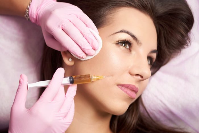 A person with long brown hair is lying down while receiving a facial cosmetic injection. The individual administering the injection, highlighting the latest cosmetic surgery trends, is wearing pink gloves and holding a syringe filled with a yellowish liquid, as well as a cotton pad near the recipient's cheek.