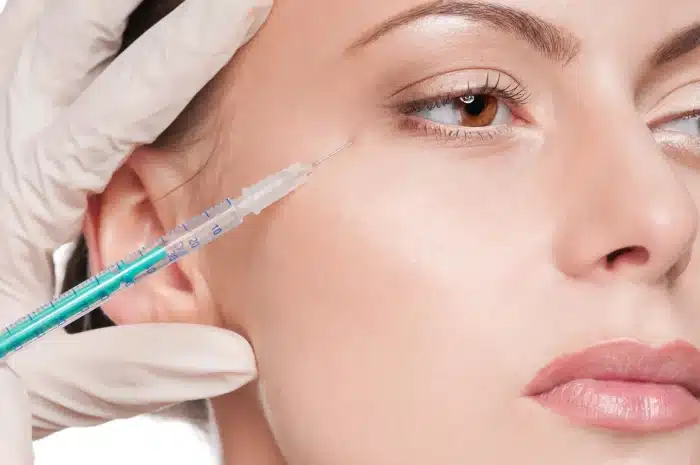 Close-up of a woman's face with a medical professional's gloved hands holding a syringe near her right eye. The woman has light skin and a neutral expression. The focus is on the syringe targeting the delicate skin at the outer corner of her eye.