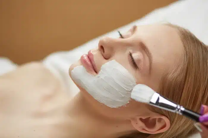 A person is lying down with their eyes closed, enjoying a facial treatment. A thick white cream is being applied to their face using a brush. The person looks calm and relaxed during the treatment.