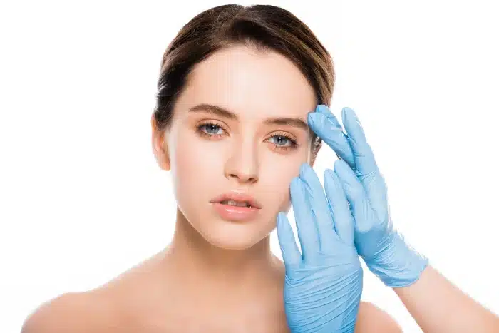 A close-up of a person with fair skin and dark hair, looking at the camera. They have a neutral facial expression while two hands in blue medical gloves gently touch their forehead and brow, suggesting a skincare or cosmetic procedure. The background is white.