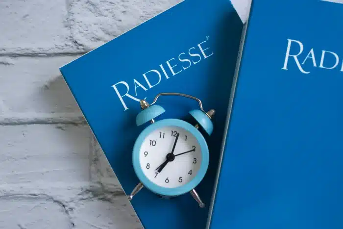 A small blue alarm clock positioned between two blue Radiesse product boxes, set against a white brick background. The clock shows 8 o'clock.