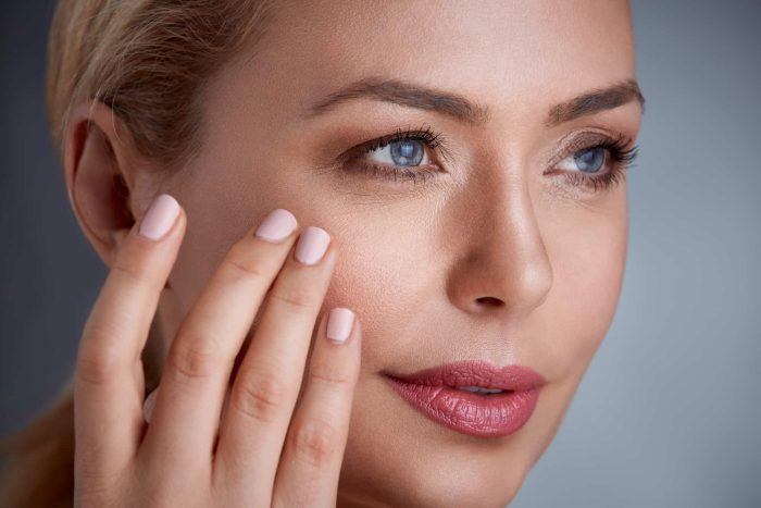 Close-up of an individual with light skin, blue eyes, and blonde hair, gently touching their cheek with neatly manicured nails. The individual has subtle makeup, with emphasis on pink lips and sleek eyebrows. The background is a soft, blurred gradient.