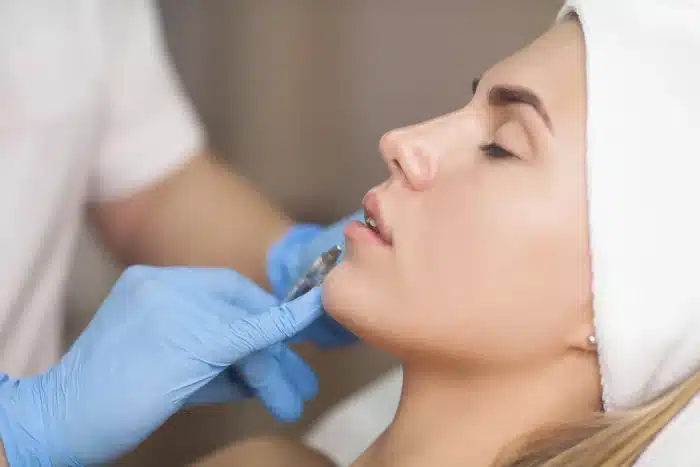 A woman is lying down with her eyes closed, wearing a white headband. A person in blue gloves is holding a syringe and administering an injection to the woman's lips. The setting appears to be a medical or spa facility.