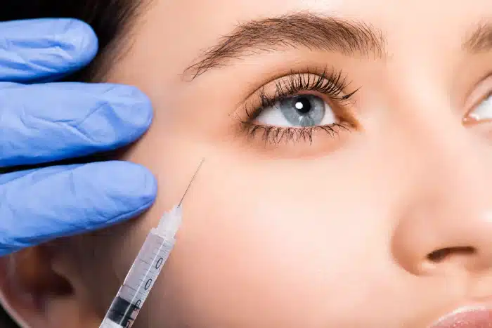 Close-up of a person receiving an injection near their eye. The person has blue eyes, long eyelashes, and well-groomed eyebrows. A gloved hand holds the side of their face while another gloved hand holds a syringe, preparing for the injection.