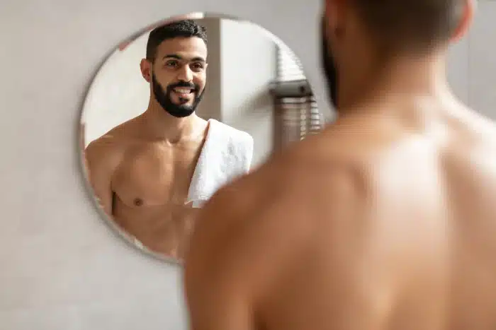 A shirtless man with a towel over his shoulder smiles while looking at himself in a round bathroom mirror. The background is minimalist with soft lighting.
