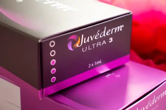A close-up image of a Juvéderm Ultra 3 product box. The black and purple packaging with the Juvéderm logo is prominently displayed, along with the product details "2 x 1mL." The background has a blurred red and pink hue.