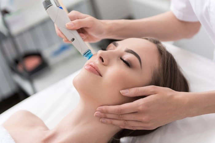 A woman is receiving a facial treatment while lying on a table with her eyes closed. A professional is using a handheld device on the woman's cheek, with their other hand gently supporting her head. The environment appears to be a spa or clinic.