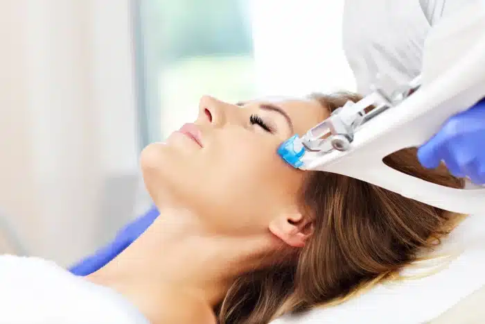 A person is lying down and receiving a cosmetic treatment on their face. A healthcare professional, wearing blue gloves, is using a handheld device near the person's temple. The scene appears to be in a clinical or spa setting.