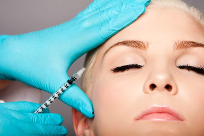 A close-up of a person with closed eyes receiving an injection on their forehead. A medical professional wearing blue gloves is administering the injection using a syringe. The person being treated appears calm and relaxed.