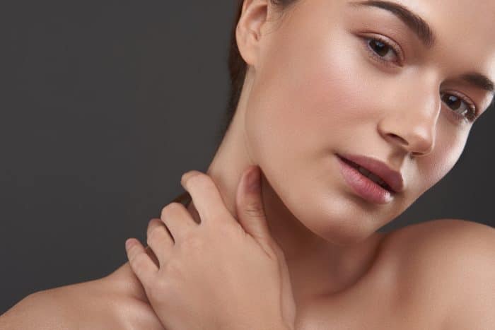 Close-up of a young person with smooth skin and minimal makeup, touching their neck with one hand. The background is dark gray, highlighting the person's natural complexion and subtle glow. The expression is calm and serene.