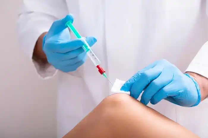 A healthcare professional, wearing blue gloves and a white coat, administers an injection into a patient's knee. The professional holds a syringe in one hand and uses a piece of gauze on the patient's skin with the other hand.