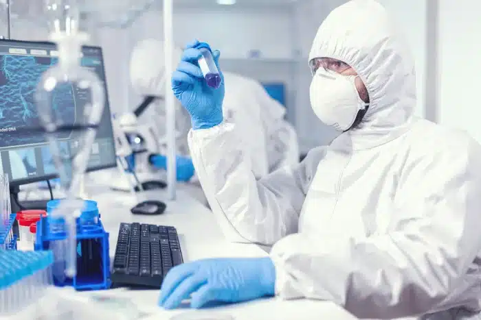 A scientist in full protective gear, including a face mask and gloves, examines a test tube containing a dark liquid in a laboratory. Nearby, there are a computer monitor displaying data, laboratory glassware, and other scientific instruments.