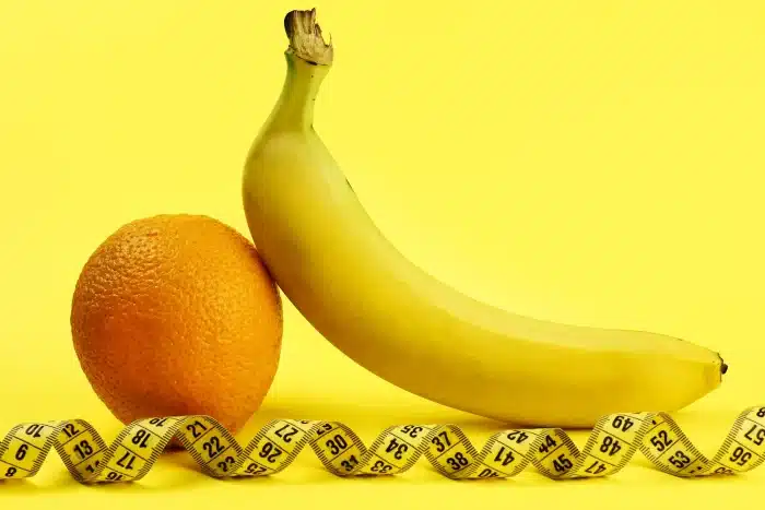 A banana and an orange are placed next to each other on a yellow background. A yellow measuring tape is laid in front of the fruits, adding a sense of health and measurement to the composition.
