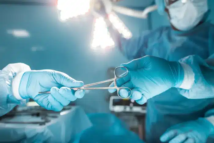 A surgical team in a brightly lit operating room, with one person passing a pair of surgical scissors to another. Both are wearing protective surgical gloves, gowns, and masks. The background shows illuminated overhead lights and surgical equipment.