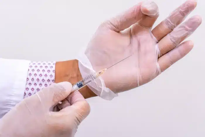 A person wearing a white coat with a patterned cuff and transparent gloves holds a syringe, about to administer an injection.