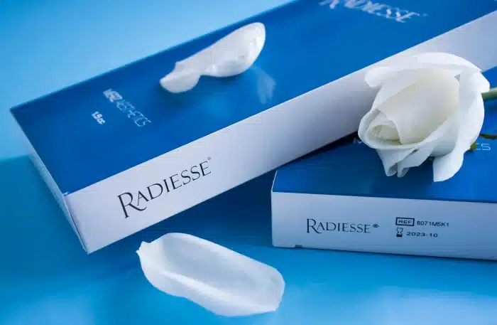 A white rose and petal rest on two blue and white boxes labeled "Radiesse" against a light blue background. One box is partially opened with a visible expiration date of 2023-10.