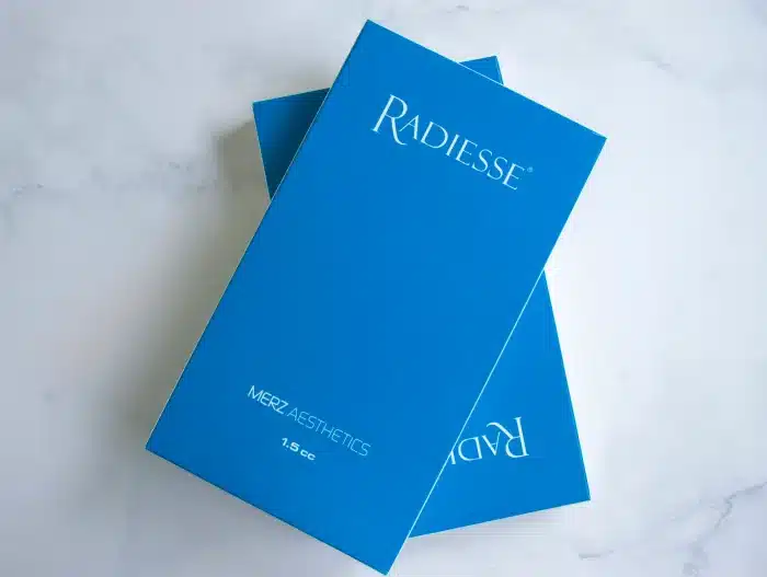 A blue rectangular box with the word "Radiesse" written on it in white font. Below that, it states "Merz Aesthetics" and "1.5 cc." Another blue box is partially visible underneath it on a white marble surface.