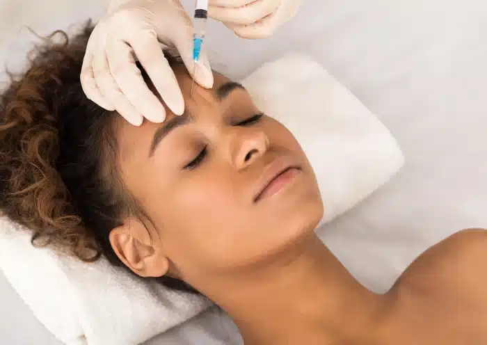 A woman with curly hair lies on a white towel while receiving a cosmetic injection in her forehead from a professional wearing white gloves. The woman has her eyes closed and appears to be relaxed.