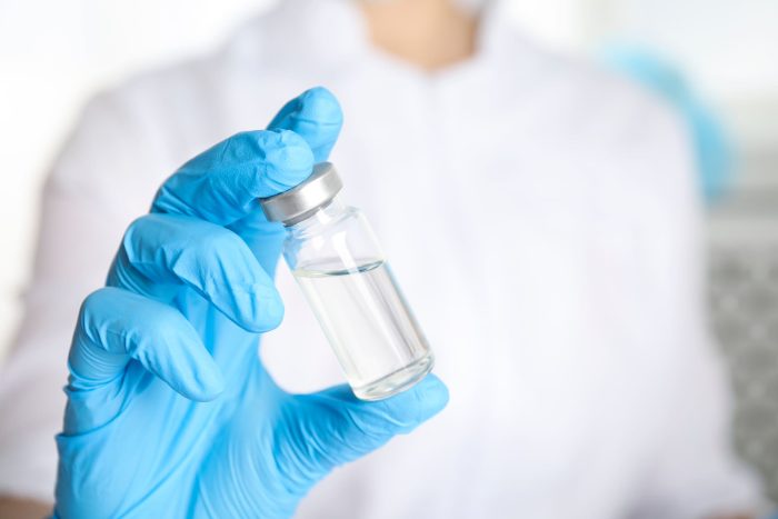 A person wearing a white lab coat and blue gloves holds a small glass vial containing a clear liquid. The background is blurred, focusing attention on the vial.