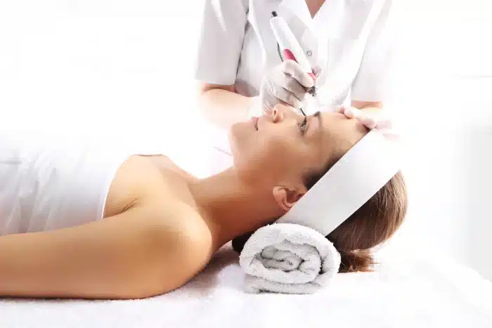 A woman is lying down with her head resting on a rolled towel. She is receiving a facial treatment from a professional wearing white attire and gloves, who is using a handheld device near her forehead. The setting appears to be a spa or wellness center.
