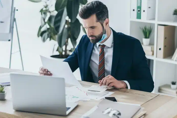 A man in a suit sits at a desk in a modern office, looking at documents. He has a disposable mask hanging below his chin. A laptop and papers are on the desk. A large plant and office supplies are visible in the background.
