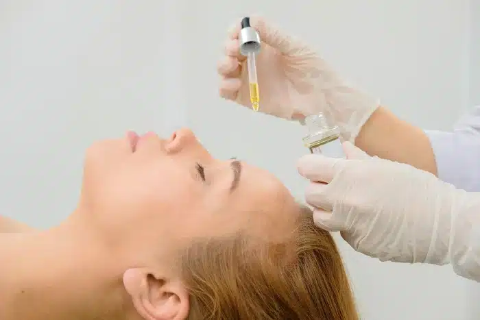 A person receives a skincare treatment lying on their back with their eyes closed. An esthetician wearing gloves uses a dropper to apply a serum or oil to the person's forehead. The background is white and out of focus.