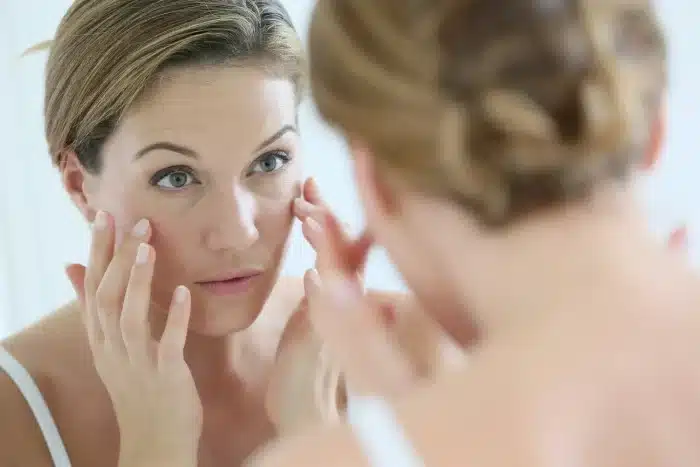 A woman with light skin and short blonde hair is standing in front of a mirror, gently touching the skin under her eyes with both hands. She appears to be examining her face closely. She is wearing a white top and the background is blurred.