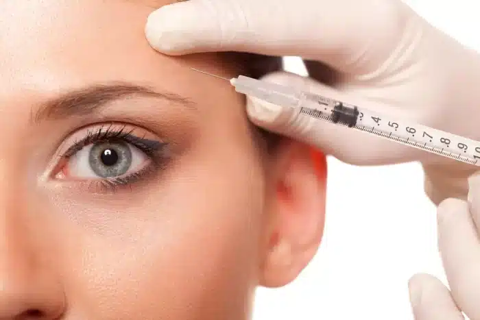 A close-up of a woman's face with a focus on her eye and forehead. A gloved hand is holding a syringe near her forehead, indicating an injection procedure, possibly for cosmetic purposes.