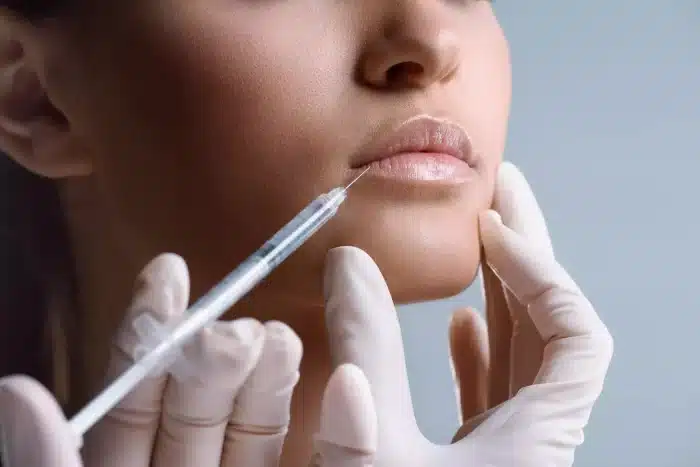 Close-up of a person receiving a lip injection. The person's face is angled to the side and their lips are slightly parted. Gloved hands are holding a syringe that is being inserted into the bottom lip, with a neutral-toned background.