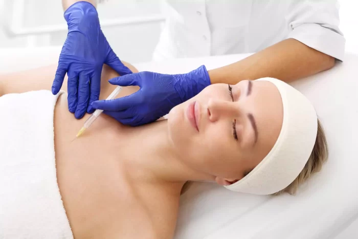 A person with a headband lies down with their eyes closed, smiling slightly. Another individual in a white coat and blue gloves administers an injection into the person's shoulder. The setting appears to be a medical or cosmetic treatment room.