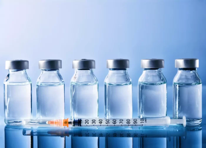 Six clear vials with silver caps, each containing a transparent liquid, are lined up on a reflective surface against a blue background. A filled syringe with an orange safety cap lies horizontally in front of the vials.