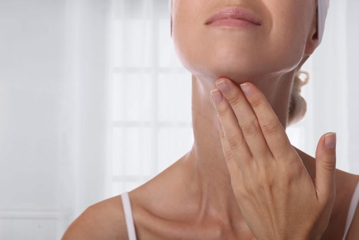 Close-up of a person's lower face, neck, and hand. The individual is gently touching their neck with their fingers, and their head is slightly tilted back. The background shows a softly lit, bright room with large windows.