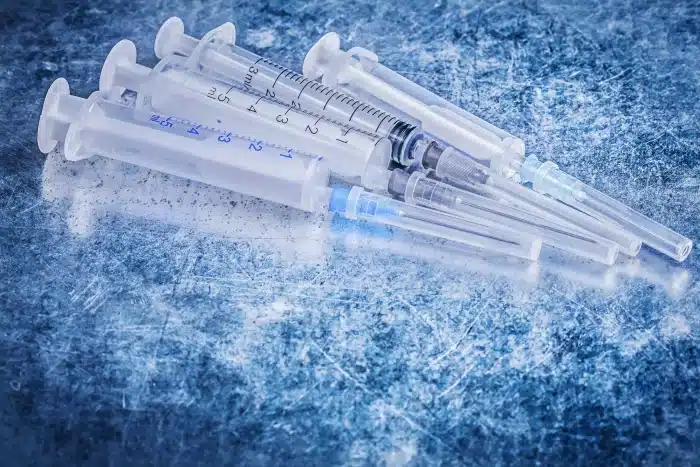 A group of medical syringes with attached needles lies on a scratched, blue-toned surface. The syringes are mostly transparent, and some have graduated markings along the sides. The background appears to be an icy or metallic texture.