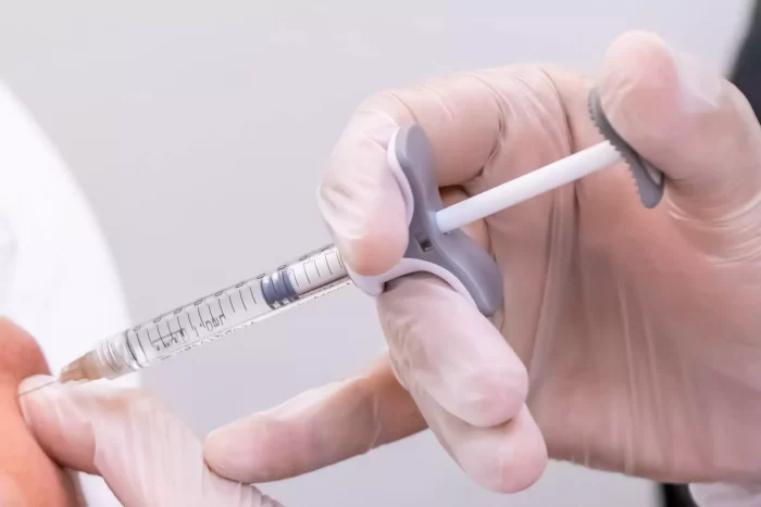Close-up of a gloved hand administering a vaccine with a syringe into a person's arm. The clear syringe is partially filled with liquid, and the person’s skin is slightly pressed around the injection site. The background is blurred and out of focus.
