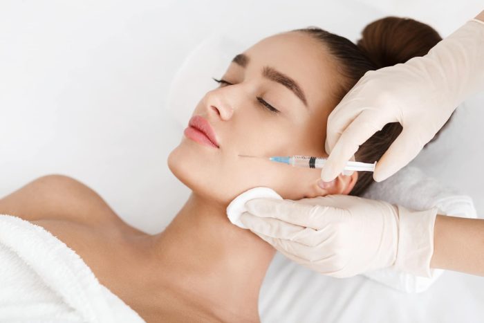 A person with closed eyes receives a facial injection administered by a professional wearing gloves. The person's head is supported with a cloth, and a cotton pad is held against their face during the procedure.