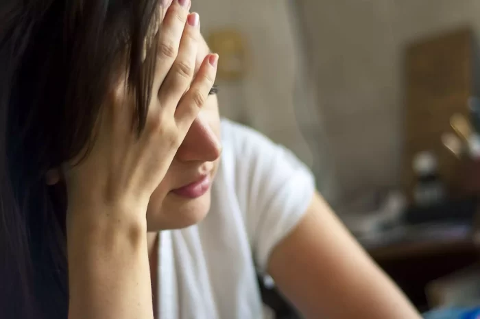 A person with long dark hair and a white shirt is sitting with their hand covering their face, appearing distressed or tired. The background is blurred, focusing attention on the person's gesture and expression.