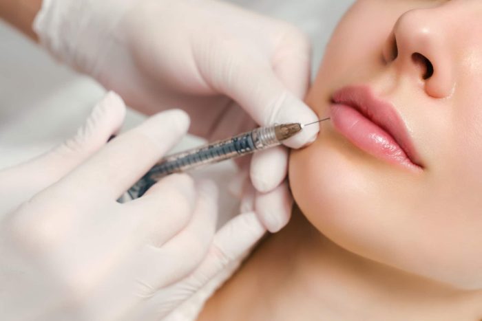 A close-up of a person receiving a Restylane injection. The person's face is partially visible, focusing on the lips. A gloved hand is holding a syringe near the person's lip, administering the Restylane Vital Light treatment. The background is blurred, highlighting the procedure and its effects.