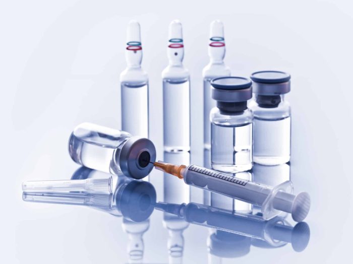 A syringe filled with liquid is positioned in front of two small, sealed vials and three ampoules containing clear liquid, possibly related to Restylane Vital Light. The background is a reflective surface, creating a mirrored effect of the medical supplies.