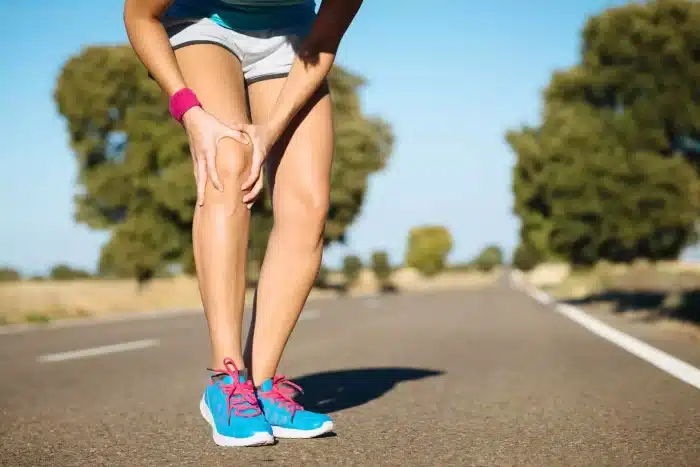 A person in athletic clothing and blue running shoes is standing on an empty road, bending forward and holding their knee as if in pain or discomfort. Trees and a clear sky can be seen in the background.
