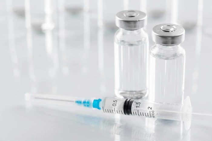 Two clear glass vials with metal caps are positioned beside a syringe filled with a clear liquid, possibly raising the question: what is Restylane? The syringe has a blue needle cap and measurement markings on its body. The background consists of more blurred vials.