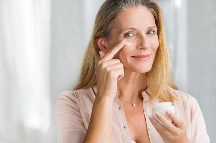 A woman with long blonde hair is smiling while applying face cream from a small jar to her cheek. She is wearing a light pink shirt and standing against a soft, blurred background.