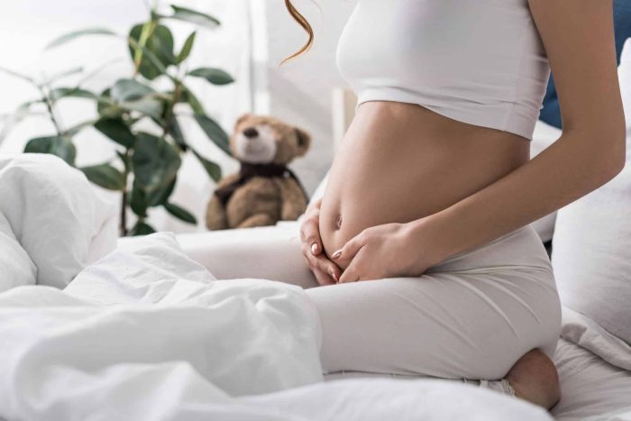 A pregnant woman dressed in a white outfit sits on a bed with her hands gently resting on her belly, contemplating the changes childbirth will bring. A teddy bear and a potted plant are visible in the background, giving a cozy and serene ambiance to the room.