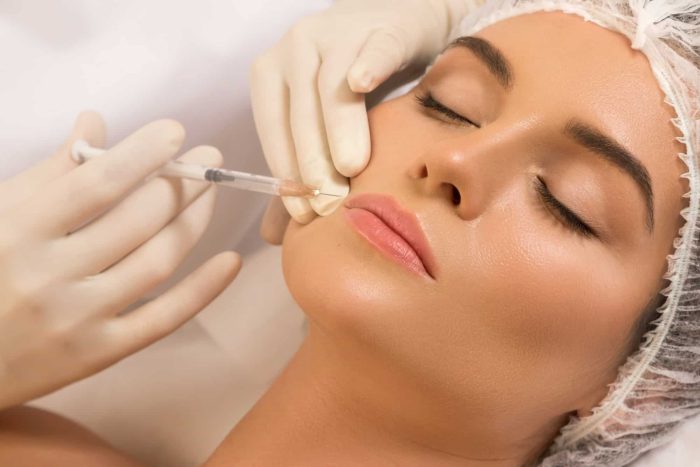 A person with closed eyes is receiving a facial injection from someone wearing gloves. The person's hair is covered with a net. The scene suggests a cosmetic or medical procedure being performed on the person's face, specifically around the lips.