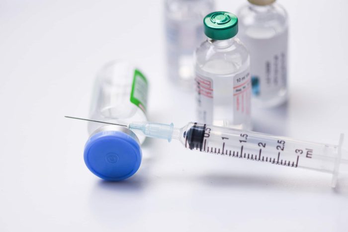 A syringe filled with liquid lies on a table next to a vial with a blue cap and several other vials with various colored caps. The liquid-filled syringe and vials are used for medication or vaccination purposes. The background is white and softly blurred.